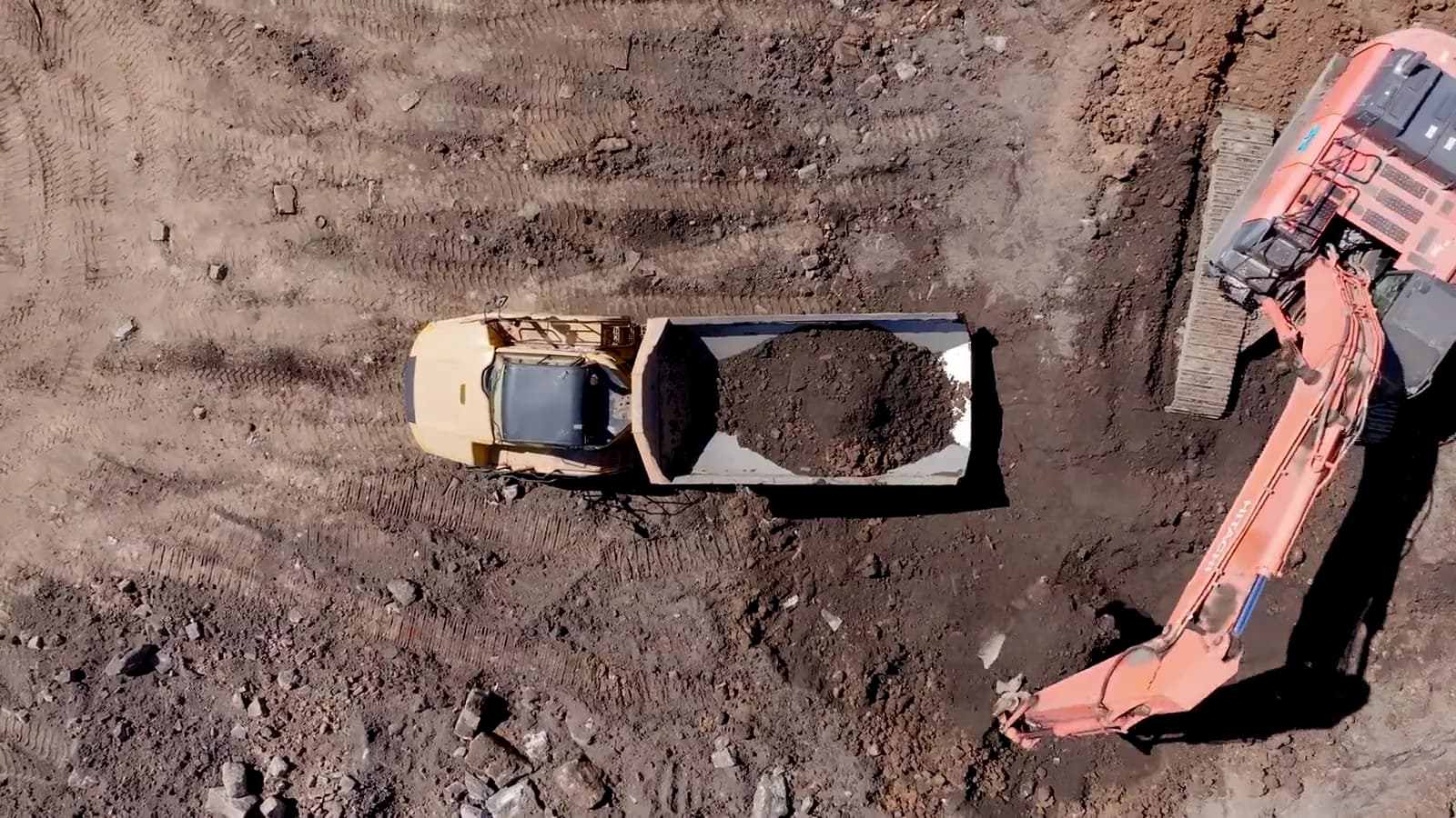 A drone looking straight down on a tracked excavator loading a tipper truck on churned ground.